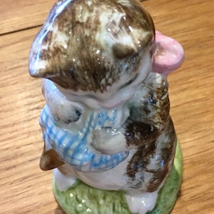 Könnte beinhalten: Eine Keramikfigur einer Katze mit braun-weißem Fell, die einen blau-weiß karierten Schal trägt. Sie hält einen rosa Gegenstand und steht auf einem grünen Sockel. Die Figur steht auf einer Holzoberfläche.