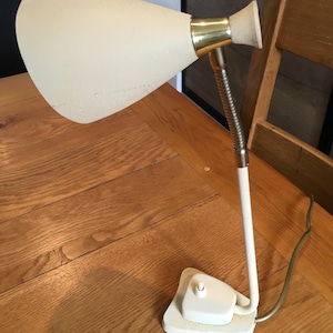 Puede incluir: Una lámpara de escritorio vintage con una pantalla blanca texturizada y un cuello color latón. La lámpara tiene una base blanca con un interruptor y un brazo flexible plateado. La lámpara está sobre una mesa de madera.