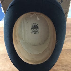 May include: A black top hat with a wide brim, viewed from the inside. The interior features a cream-colored lining and a label with text. The hat is likely vintage, with signs of wear.