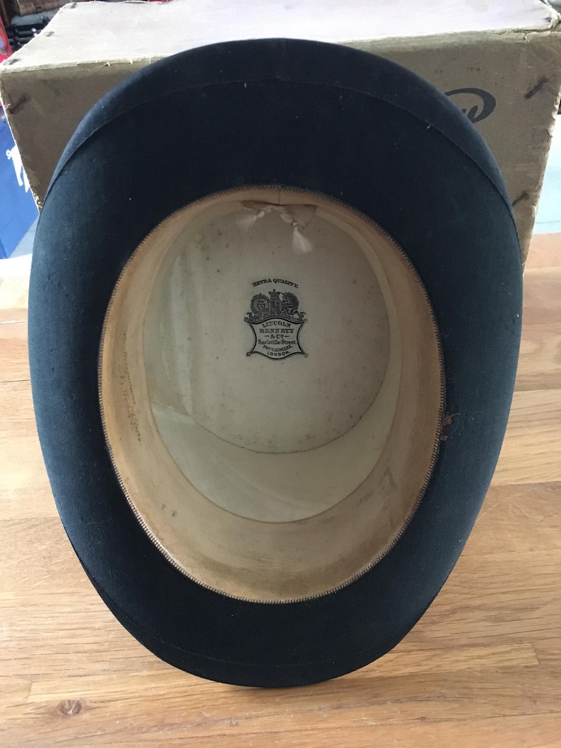 May include: A black top hat with a wide brim, viewed from the inside. The interior features a cream-colored lining and a label with text. The hat is likely vintage, with signs of wear.