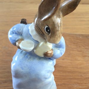 May include: A porcelain figurine of a brown rabbit wearing a blue dress and holding a white cup. The rabbit has brown ears and a white collar.