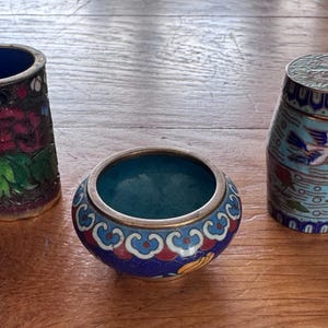 May include: Three cloisonné enamel vessels including a tall cylindrical vase with a pink floral design, a small round bowl with blue and red scalloped patterns, and a lidded jar featuring a light blue background with a bird and floral motif.