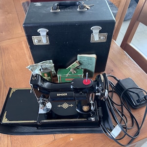 Könnte beinhalten: Vintage schwarze Singer Nähmaschine mit goldenen Akzenten, einer schwarzen Tragetasche und einem Fußpedal. Die Maschine hat einen roten Spulenhalter. Zubehör: eine grüne Box, eine Dose und ein Handbuch.