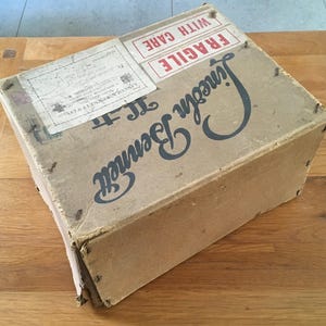 May include: A vintage cardboard box with the name "Lincoln Bennett" in large, dark blue script. The box has a "Fragile, With Care" label and other shipping labels. The box is secured with small metal staples and shows signs of wear.