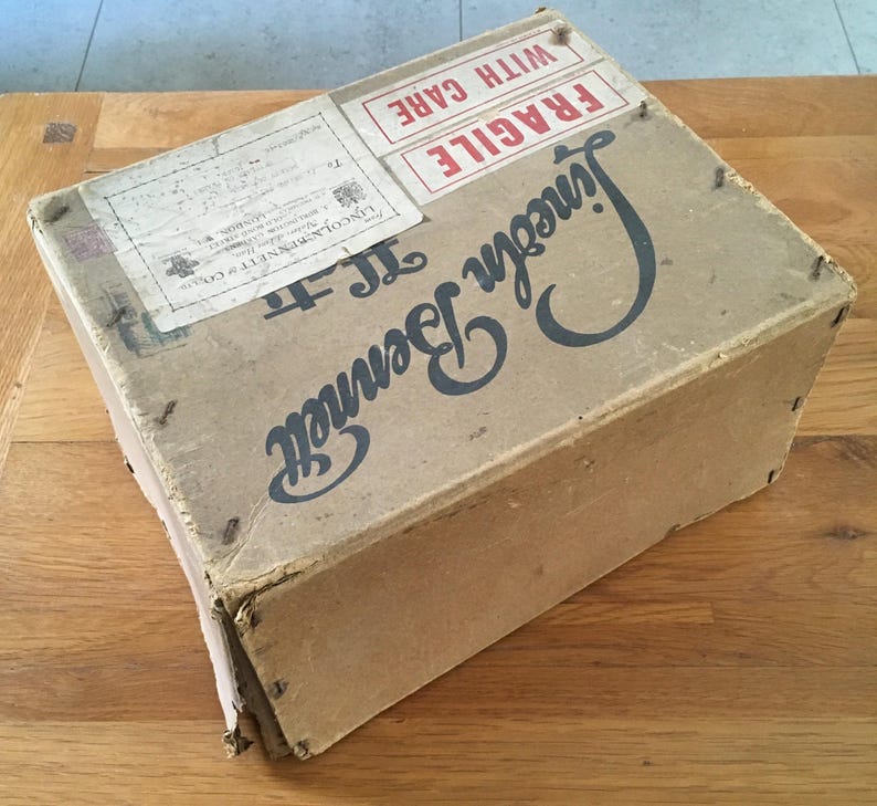 May include: A vintage cardboard box with the name "Lincoln Bennett" in large, dark blue script. The box has a "Fragile, With Care" label and other shipping labels. The box is secured with small metal staples and shows signs of wear.
