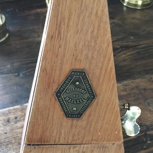 Può includere: Un metronomo in legno di forma trapezoidale. Il metronomo ha una placca metallica con la scritta "METRONOME DE MAELZEL" e le città di Parigi, Londra, Bruxelles e Olanda. Una chiave è visibile sul lato.