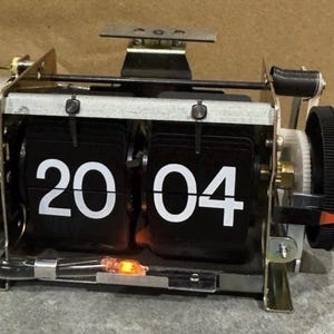 May include: Vintage flip clock displaying "20 04" in white numerals on black panels. The clock has a metal frame with a black dial for setting the time. A small orange light is visible.