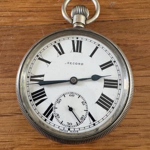 Op de afbeelding: Een zilveren zakhorloge met een witte wijzerplaat en zwarte Romeinse cijfers. Het horloge heeft een kleine secondenwijzer en het woord "RECORD" staat op de wijzerplaat. Het horloge is gemarkeerd met "SWISS MADE".