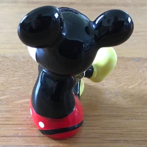 May include: A glossy figurine of Mickey Mouse, viewed from the back. The figure has large black ears, a black body, red shorts with white buttons, and yellow gloves. The figurine is on a wooden surface.