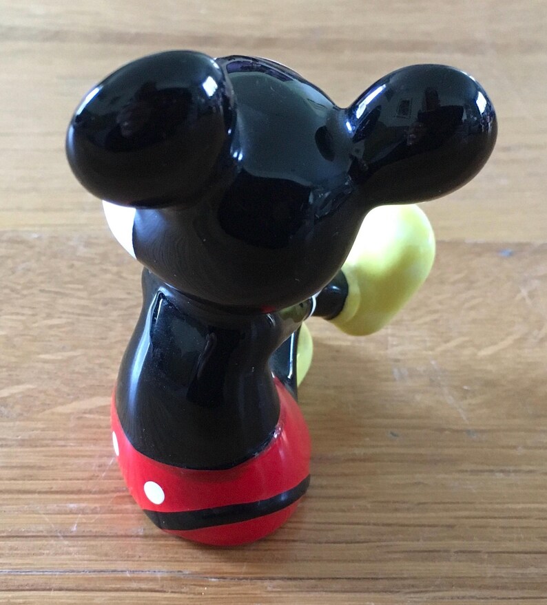 May include: A glossy figurine of Mickey Mouse, viewed from the back. The figure has large black ears, a black body, red shorts with white buttons, and yellow gloves. The figurine is on a wooden surface.