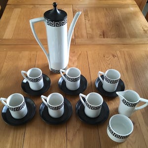 May include: A white and black ceramic coffee set featuring a Greek key pattern. The set comprises a tall coffee pot, six cups with saucers, a creamer, and a sugar bowl. The coffee pot has a black lid.