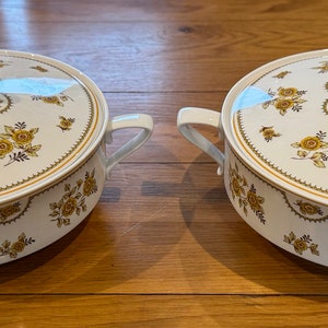 Może przedstawiać: Dwa białe ceramiczne naczynia do serwowania z pokrywkami. Naczynia mają kwiatowy wzór z żółtymi i brązowymi kwiatami na białym tle.