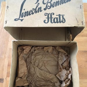 May include: An open cardboard box with the text "Lincoln Bennett Hats" printed on the lid. The box is beige and contains crumpled brown packing paper. The box sits on a wooden surface.