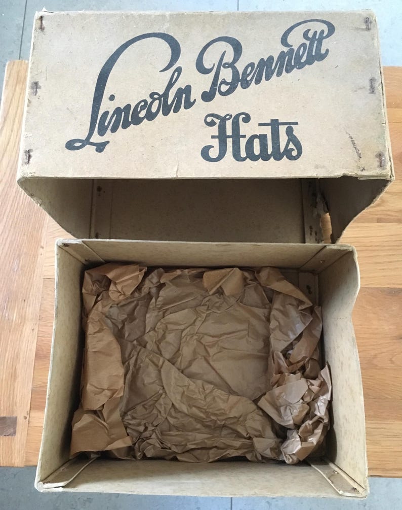May include: An open cardboard box with the text "Lincoln Bennett Hats" printed on the lid. The box is beige and contains crumpled brown packing paper. The box sits on a wooden surface.