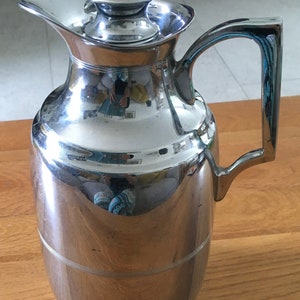 Può includere: Una caraffa termica in metallo argentato lucido con un coperchio e un manico. La caraffa ha un corpo arrotondato e un collo stretto.