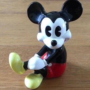 May include: A ceramic Mickey Mouse figurine in a seated pose. The figure has black ears, a black body, red shorts, white gloves, and yellow shoes. The figurine is a collectible item.
