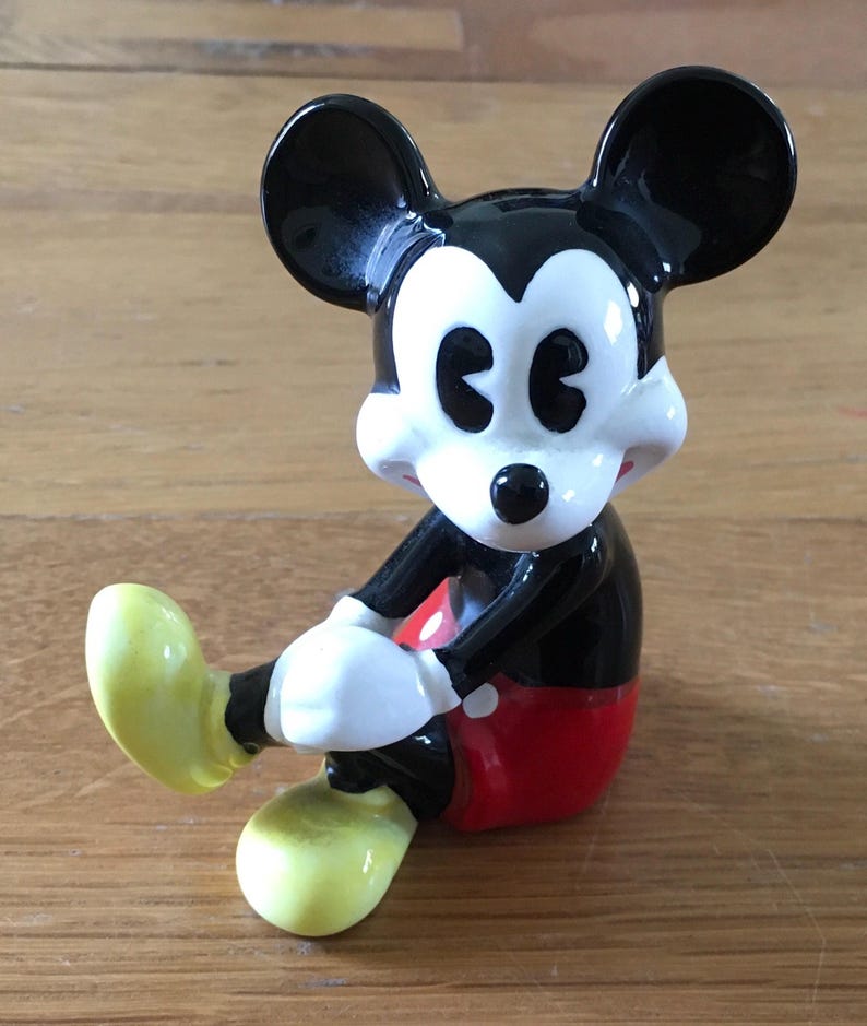 May include: A ceramic Mickey Mouse figurine in a seated pose. The figure has black ears, a black body, red shorts, white gloves, and yellow shoes. The figurine is a collectible item.