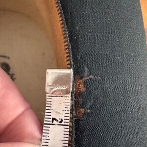 May include: Close-up of a black fabric with a zipper and a measuring tape. The tape shows a measurement of approximately 2 inches. The fabric has a small tear near the zipper.