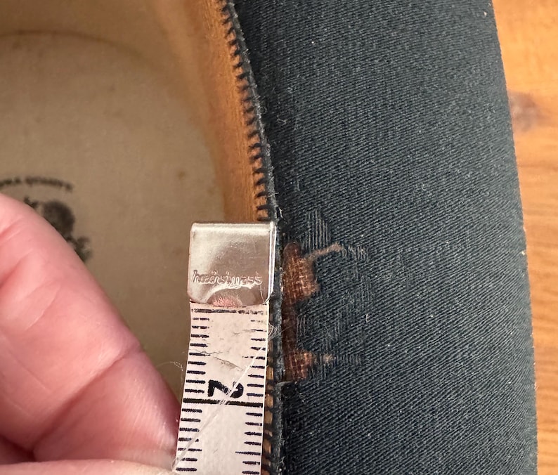 May include: Close-up of a black fabric with a zipper and a measuring tape. The tape shows a measurement of approximately 2 inches. The fabric has a small tear near the zipper.