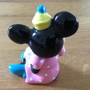 May include: A ceramic figurine of a cartoon mouse, featuring a black head with large ears, a pink dress with white polka dots, and a blue and yellow hat. The figurine is sitting down.