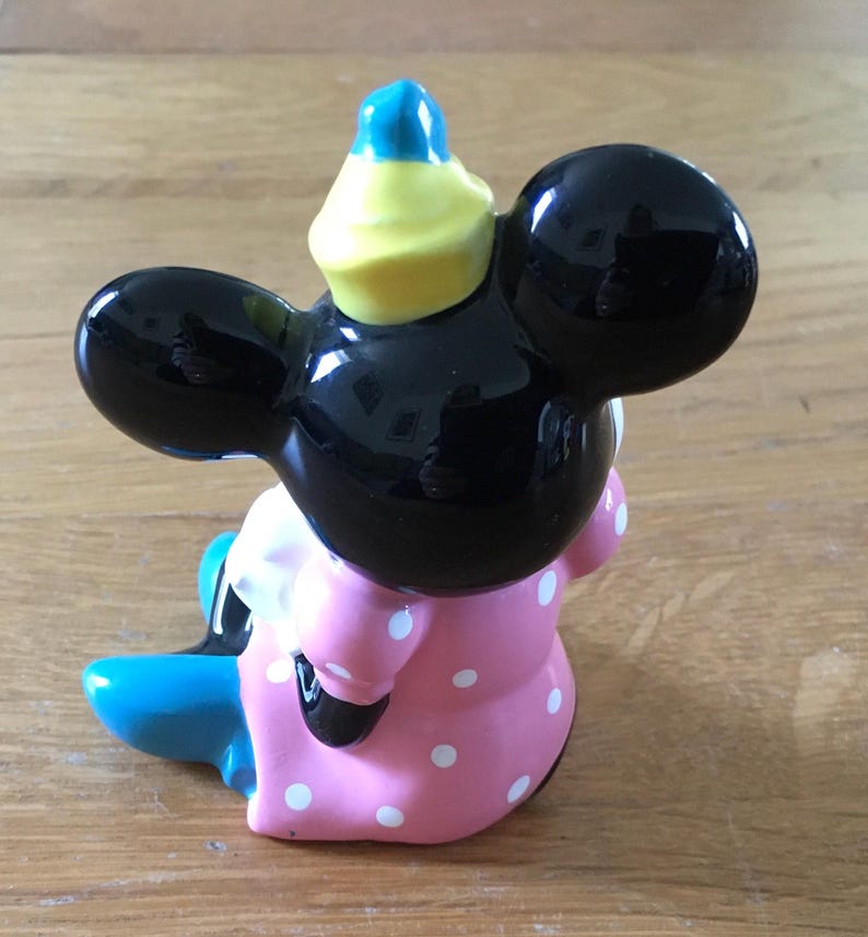 May include: A ceramic figurine of a cartoon mouse, featuring a black head with large ears, a pink dress with white polka dots, and a blue and yellow hat. The figurine is sitting down.