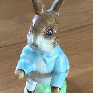May include: A ceramic rabbit figurine, dressed in a light blue jacket and white shirt. The rabbit has brown ears and a white face. It stands on a green base. This decorative figurine is a classic design.