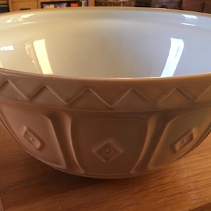 May include: A large, beige ceramic mixing bowl with a white interior and a decorative diamond pattern. The bowl has a scalloped rim and is sitting on a wooden surface.