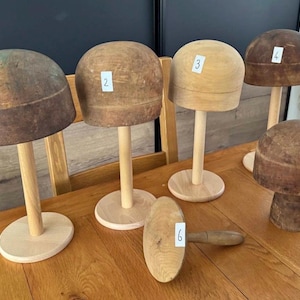 May include: A collection of wooden hat forms and a mallet on a wooden table. The hat forms are various sizes and colors, from light to dark brown, and are mounted on wooden stands. The mallet is light brown with a rounded head and a handle.