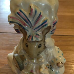 Può includere: Una statuetta in ceramica di un topo con cappello e vestito. Il topo è marrone chiaro con occhi neri e tiene in mano un piccolo oggetto rotondo. L'abito è bianco con pois blu e dettagli floreali. Il cappello ha dettagli rosa e blu.