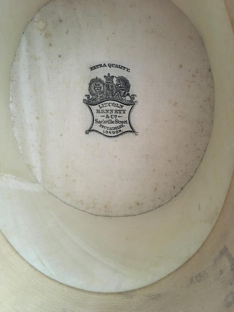 May include: Inside view of a cream-colored hat with a visible brim. The hat's interior features a faded, circular label with the text "EXTRA QUALITY" and "LINCOLN BENNETT & CO - Sackville Street, PICCADILLY, LONDON."