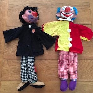 May include: Two vintage clown puppets. One puppet wears a black coat, checkered pants, and black shoes. The other puppet has a red and yellow shirt, red and white checkered pants, and purple shoes. Both puppets have painted faces.