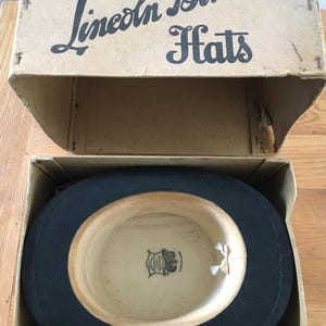 May include: A black top hat inside its original cardboard box. The box is labeled "Lincoln Bennett Hats" in black script. The hat has a cream-colored interior with a decorative emblem. The box is open, revealing the hat.