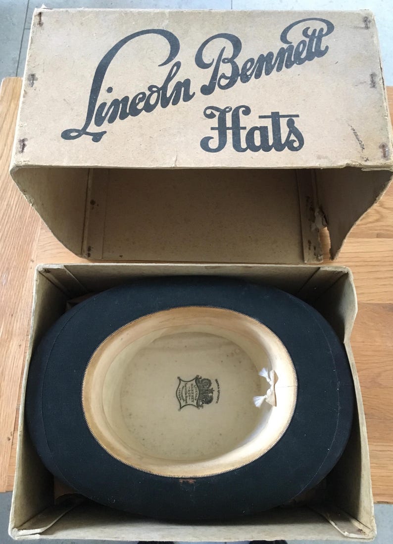 May include: A black top hat inside its original cardboard box. The box is labeled "Lincoln Bennett Hats" in black script. The hat has a cream-colored interior with a decorative emblem. The box is open, revealing the hat.