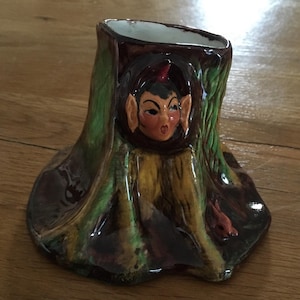 Könnte beinhalten: Eine Keramikvase in Form eines Baumstumpfes mit grüner, brauner und gelber Glasur. Ein kleiner, rothaariger Kobold schaut aus dem Hohlraum des Baumstumpfes. Ein kleines, rotes Kaninchen befindet sich am Fuß des Baumstumpfes.