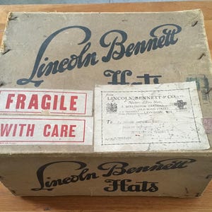May include: A vintage cardboard box with the text "Lincoln Bennett Hats" in black script. The box has "Fragile With Care" stickers and an address label. The box is tan with visible wear.