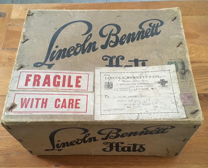 May include: A vintage cardboard box with the text "Lincoln Bennett Hats" in black script. The box has "Fragile With Care" stickers and an address label. The box is tan with visible wear.