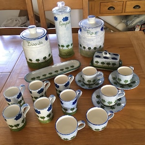 May include: A set of hand-painted ceramic kitchen items with a tree motif. The collection includes storage jars labelled "Biscuits", "Spaghetti", and "Flour", plus cups, saucers, a butter dish, and a serving platter. The colour palette is white, blue, and green.
