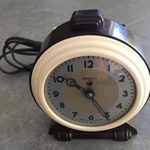 May include: A classic alarm clock with a cream-coloured face and a dark brown casing. The clock face displays black numerals and hands, with the brand name "FERRANTI". The clock has a rectangular base and a black power cord. Made in England.