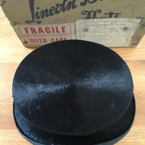 May include: A black bowler hat with a rounded crown and a flat brim. The hat is made of a dark, textured material. The hat is sitting in front of a cardboard box with the text "Lincoln & Bennett" and "Fragile with care".