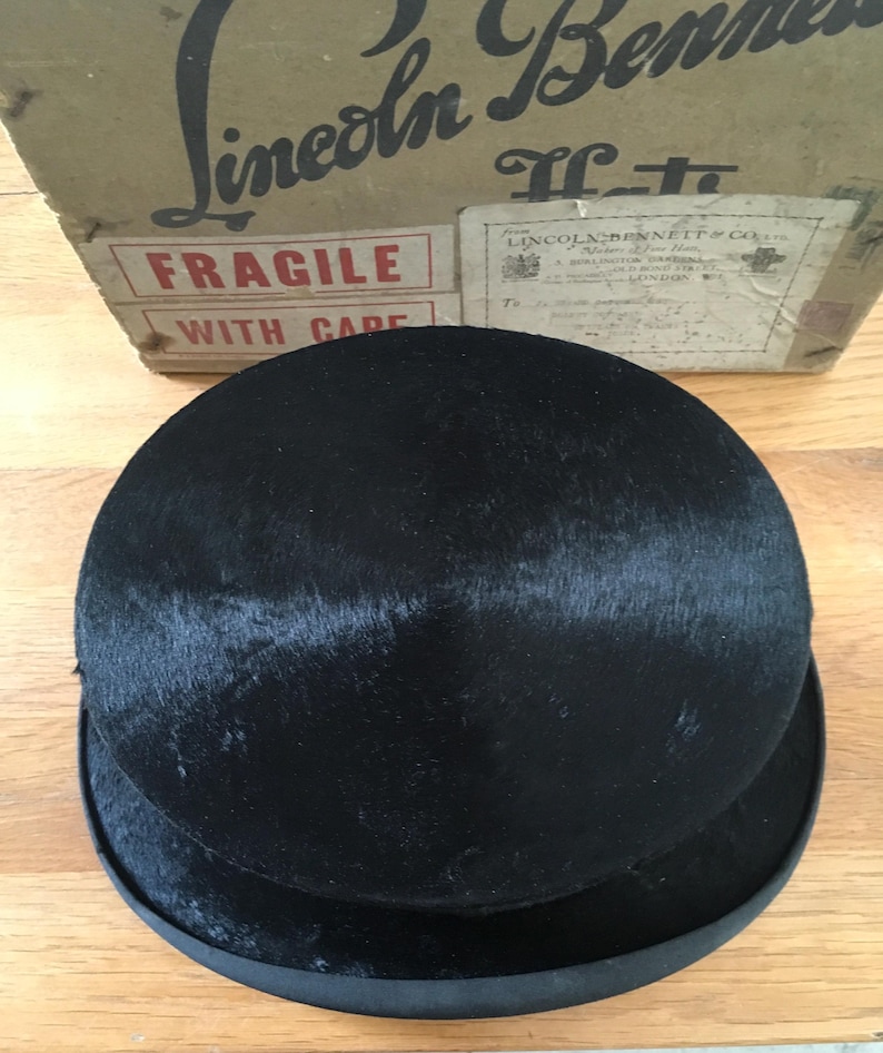 May include: A black bowler hat with a rounded crown and a flat brim. The hat is made of a dark, textured material. The hat is sitting in front of a cardboard box with the text "Lincoln & Bennett" and "Fragile with care".