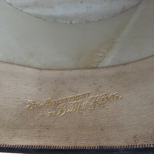 May include: Interior view of a hat, showcasing the lining and brim. The lining is off-white, and the brim is a darker color. Gold lettering reads "By Appointment to B.M. King."