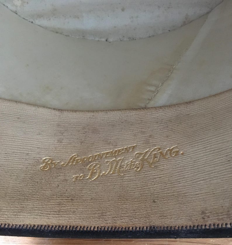 May include: Interior view of a hat, showcasing the lining and brim. The lining is off-white, and the brim is a darker color. Gold lettering reads "By Appointment to B.M. King."