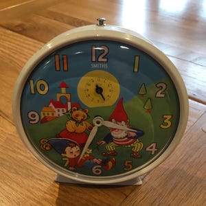 May include: A vintage Smiths alarm clock with a cream-colored metal frame. The clock face features a whimsical illustration of gnomes, a bear, and a house against a blue and green background. The clock hands are white.