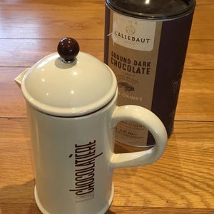 Op de afbeelding: Een crèmekleurige keramische cafetière met een bruine knop en handvat, naast een donkerbruine cilindrische container met gemalen pure chocolade. De cafetière heeft de woorden "La Chocolatiere" op de zijkant.