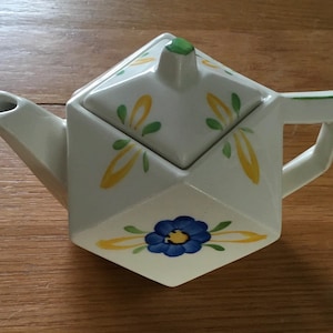 Könnte beinhalten: Weiße Keramik-Teekanne mit geometrischem Design. Die Teekanne hat eine blaue Blume und gelb-grüne Blätter. Der Deckel hat einen grünen Knopf. Der Griff ist rechteckig. Die Teekanne steht auf einer Holzoberfläche.
