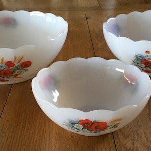 May include: Three white glass bowls with scalloped edges and floral designs. The bowls feature a cluster of red poppies, blue flowers, and wheat stalks. The bowls are arranged on a wooden surface.