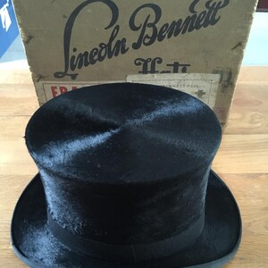 May include: A black top hat with a wide brim and a black band, sitting in front of a cardboard box. The box has the words "Lincoln Bennett" printed on it in a vintage font.