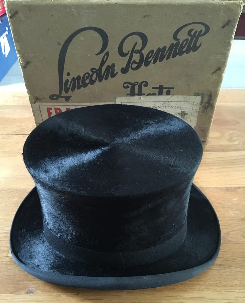 May include: A black top hat with a wide brim and a black band, sitting in front of a cardboard box. The box has the words "Lincoln Bennett" printed on it in a vintage font.