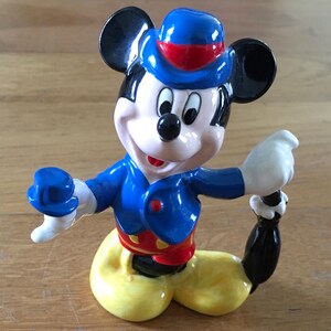 May include: A ceramic figurine of Mickey Mouse in a seated pose. Mickey is wearing a blue jacket, red shorts, yellow pants, and a blue hat with a red band. His black ears and tail are visible. The figurine has a glossy finish.