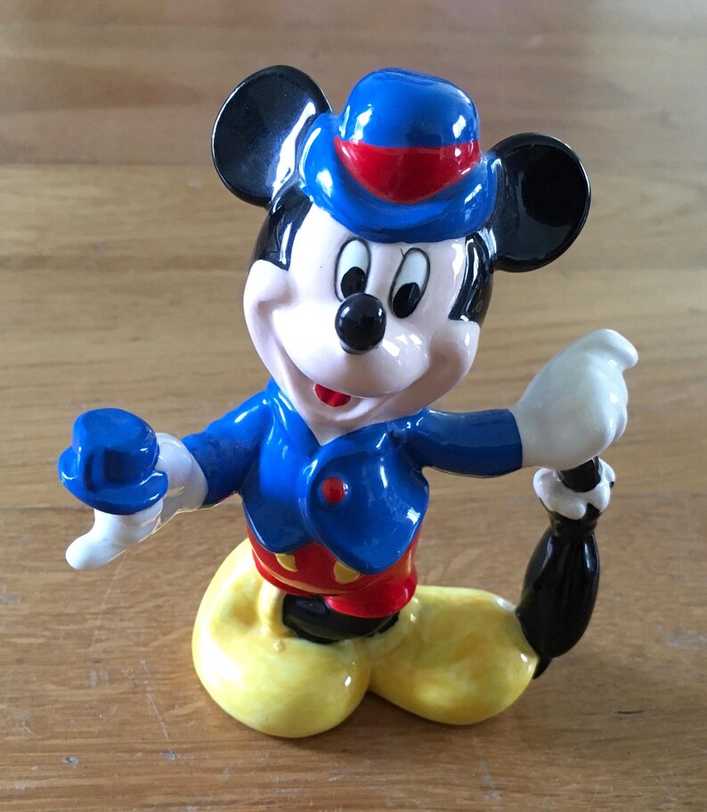 May include: A ceramic figurine of Mickey Mouse in a seated pose. Mickey is wearing a blue jacket, red shorts, yellow pants, and a blue hat with a red band. His black ears and tail are visible. The figurine has a glossy finish.
